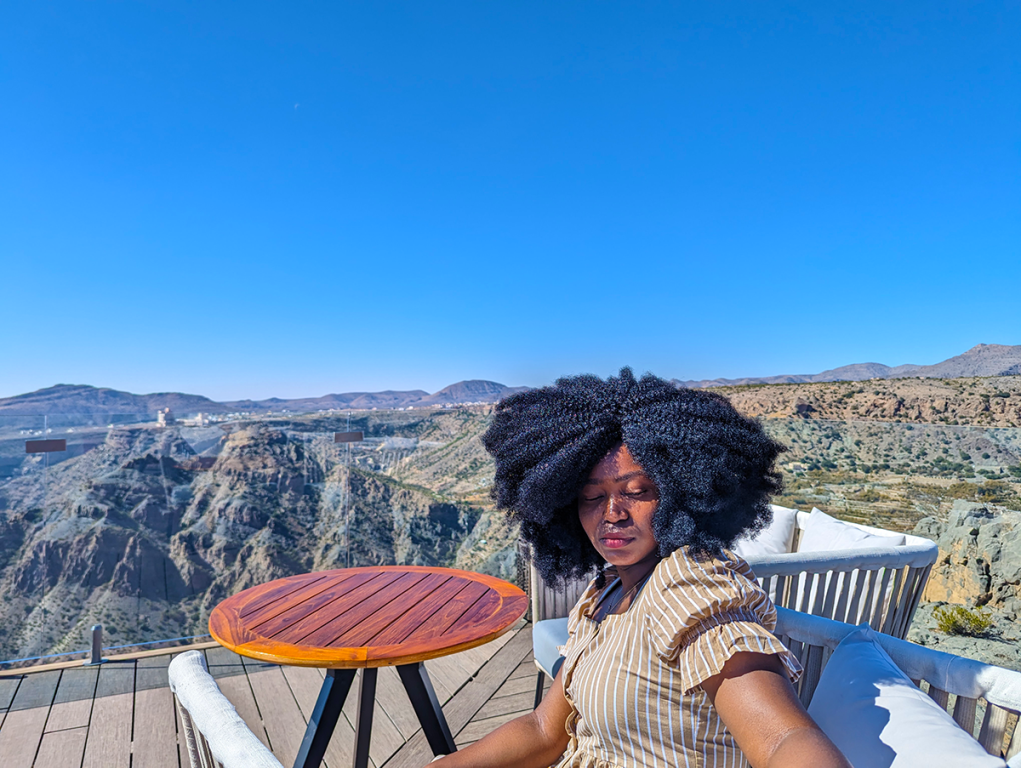 Captured from Diana's viewpoint during a visit to Oman, an image of Jadesola with afro hair sitting at a table, mesmerized by the scenic mountains.
