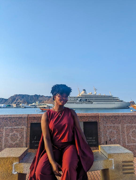 Jadesola is sitting on a bench. Behind her is the Muttrah river with a white yacht passing by.